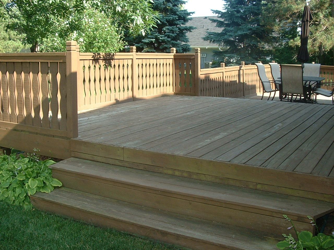 A raised, stained wood deck with railings and steps, surrounded by green trees and grass, with outdoor furniture nearby.