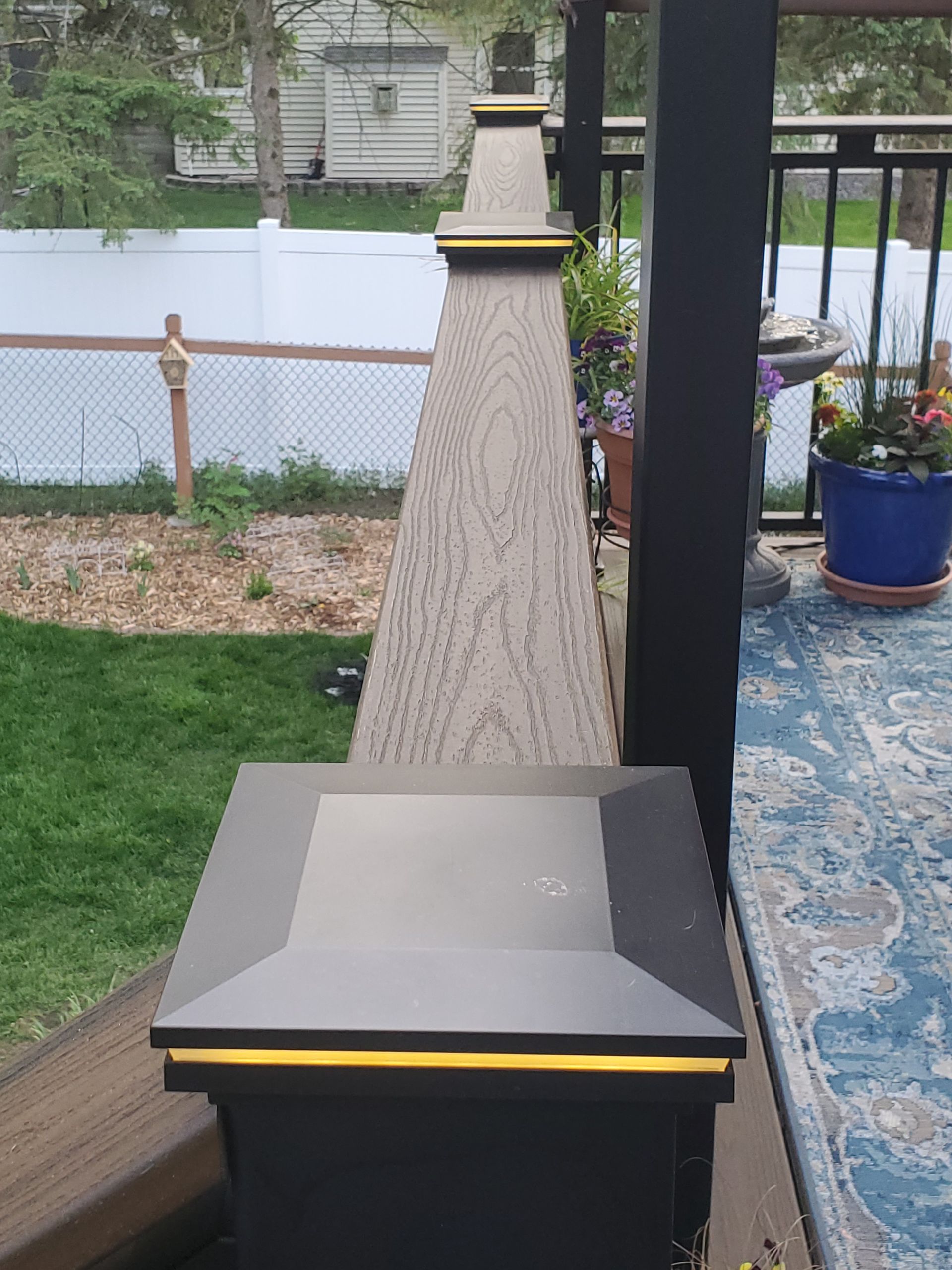 A close-up of a modern, black deck post cap with integrated lighting, overlooking a green backyard and a patio.