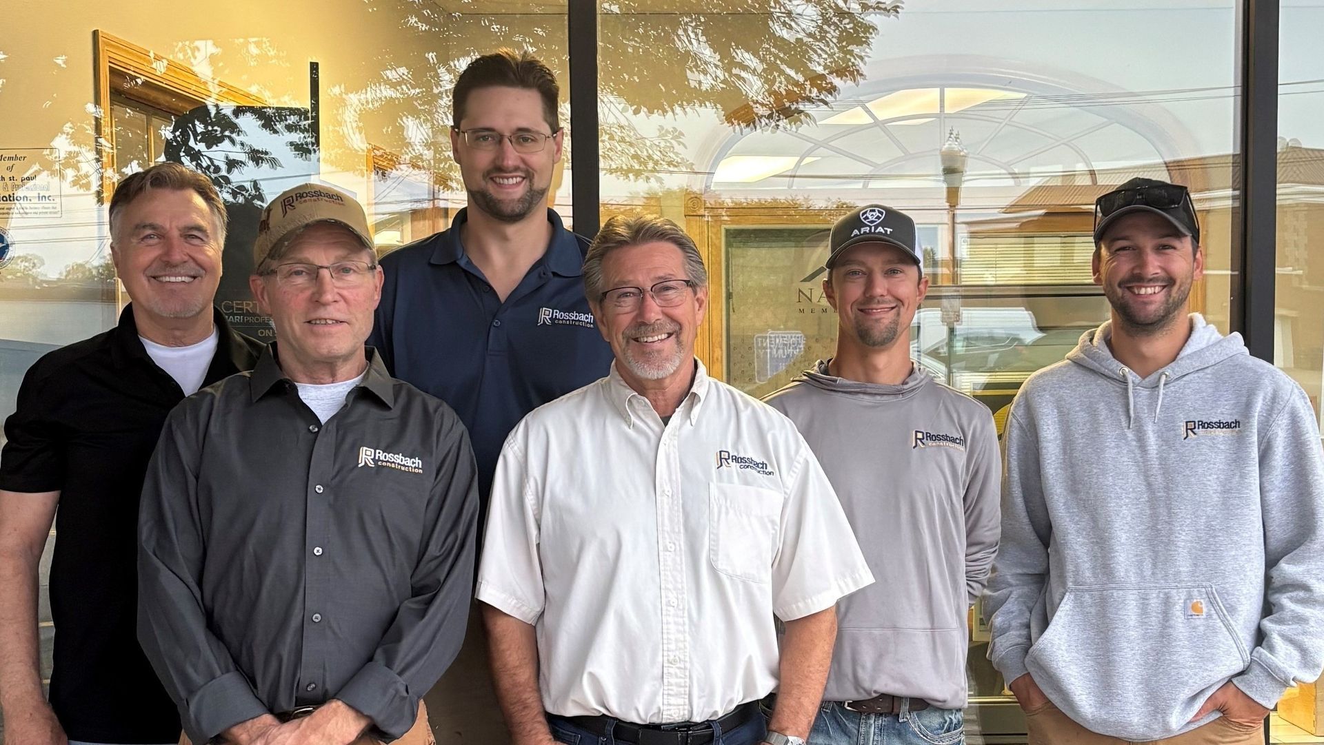 Six individuals stand in a line smiling, posing in front of a glass storefront reflecting a tree and light fixtures.