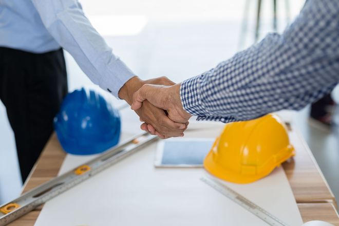 Two men shaking hands over a table with blueprints, hardhats, and tools