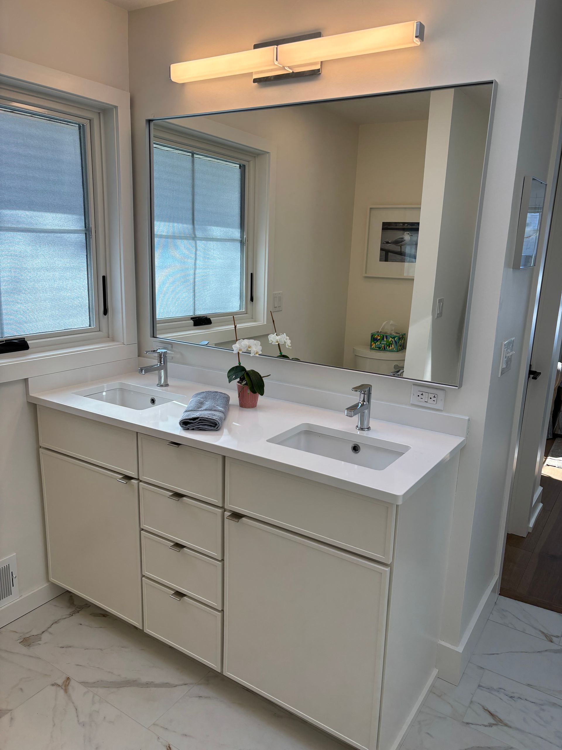 Bathroom with white double vanity, large mirror, and overhead lighting. Two sinks, window, and marble flooring.