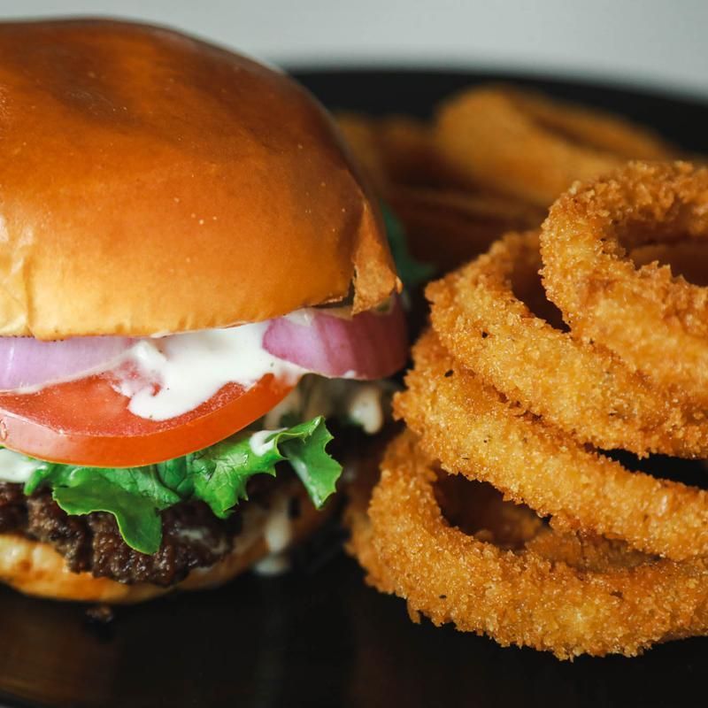 Burger with lettuce, tomato, red onion, and mayonnaise, served with onion rings on a black plate.