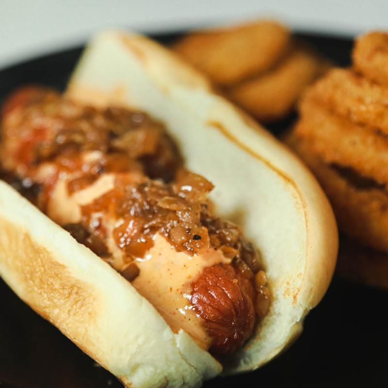 Hot dog in bun with onion topping and side of onion rings and potato wedges.