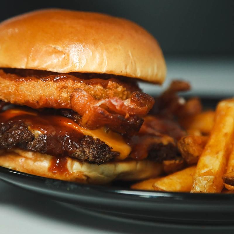 Burger with cheese, onion ring, bacon, and fries on a black plate.