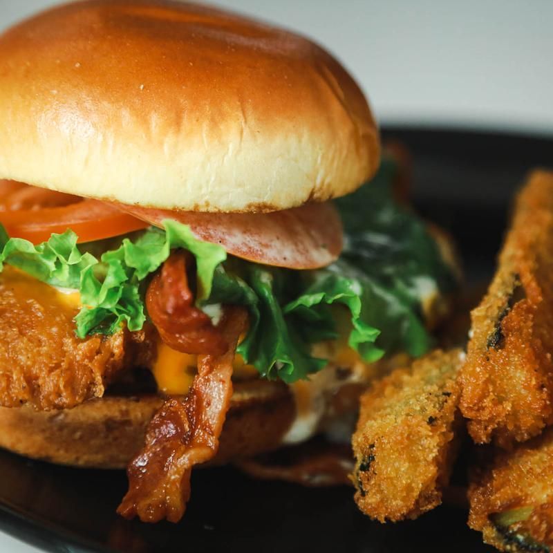 Fried chicken sandwich with bacon, tomato, lettuce, and fried zucchini on a black plate.