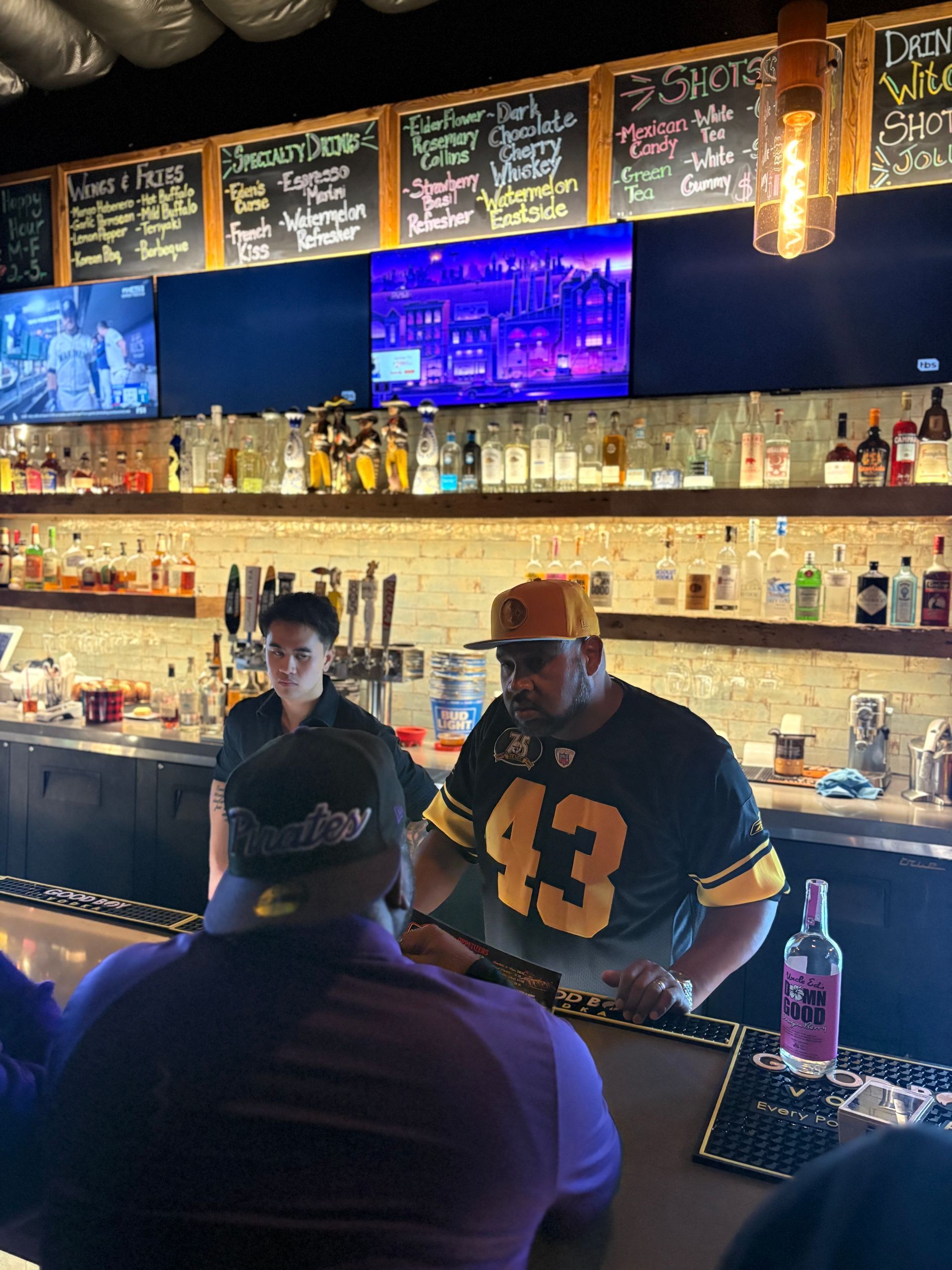 Bartender serving at a bar. Person in a sports jersey, bottles on shelves, drink menu.