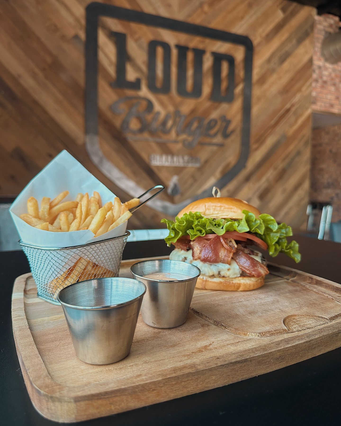 Burger and fries meal on a wooden board. The burger has bacon and lettuce. Fries are in a wire basket.