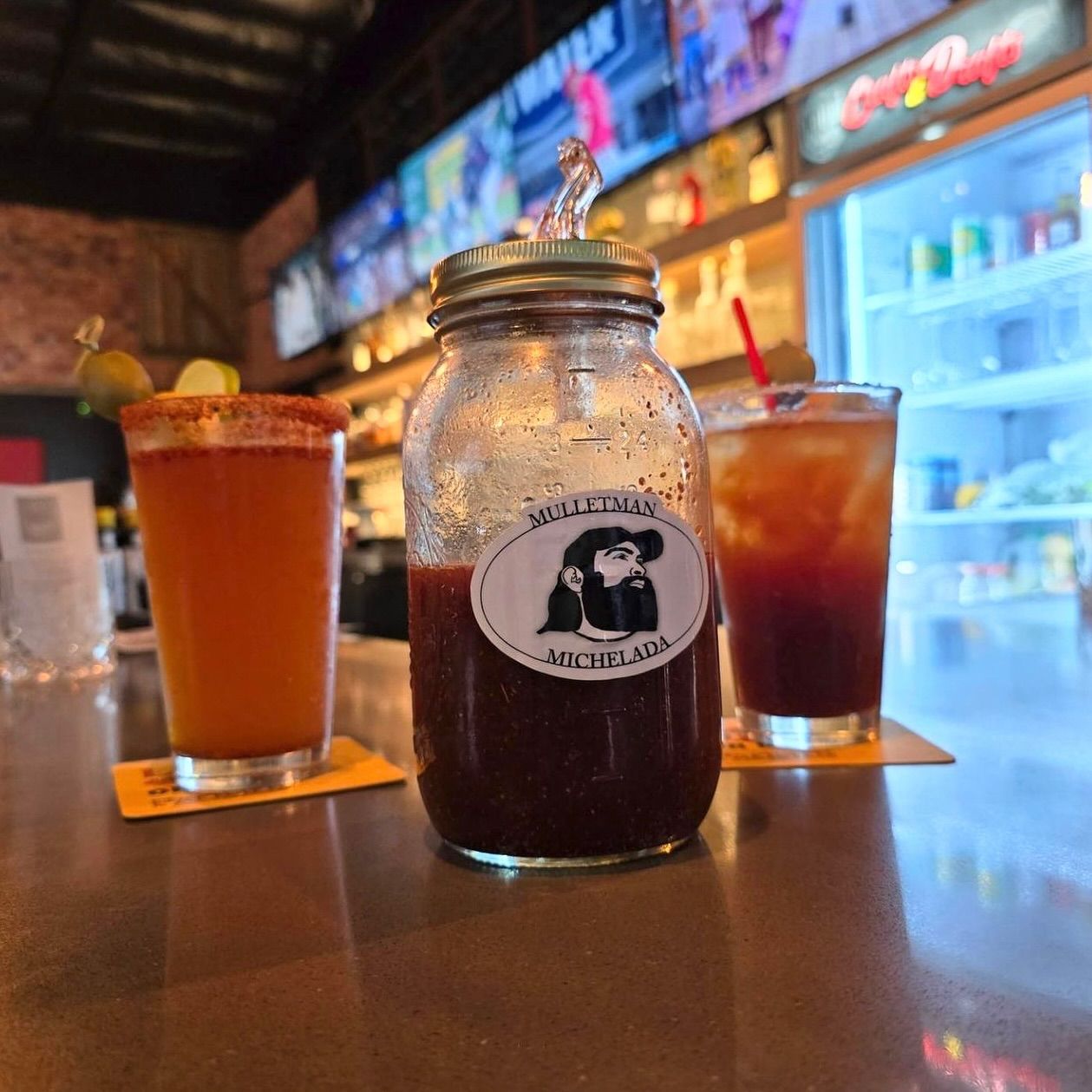 Bar scene: Jar of Bloody Mary mix and two cocktails on a bar counter. One drink has a lime wedge and red-rimmed glass.