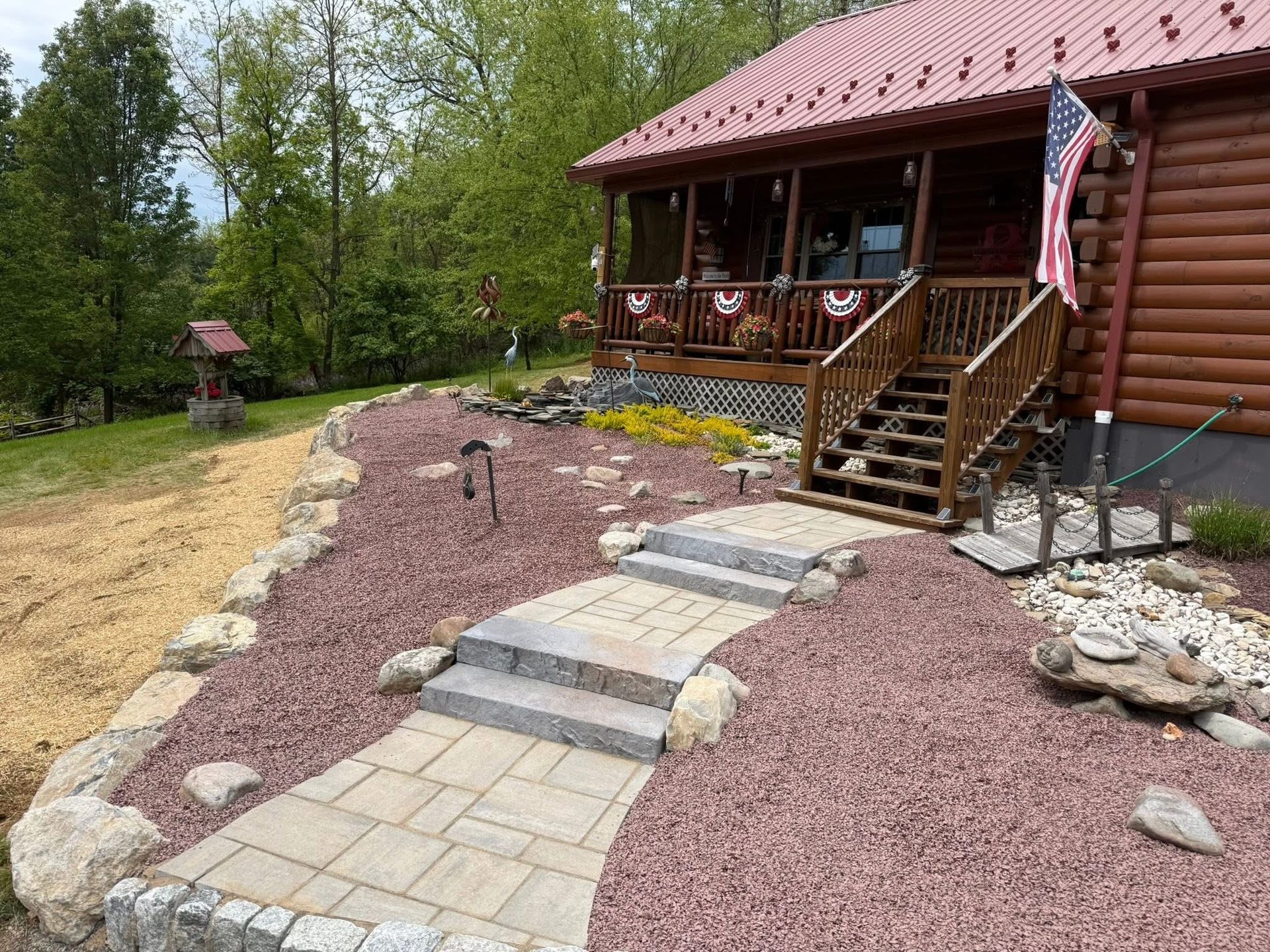 Log cabin porch with steps, paver path, and landscaped rock garden in a wooded yard