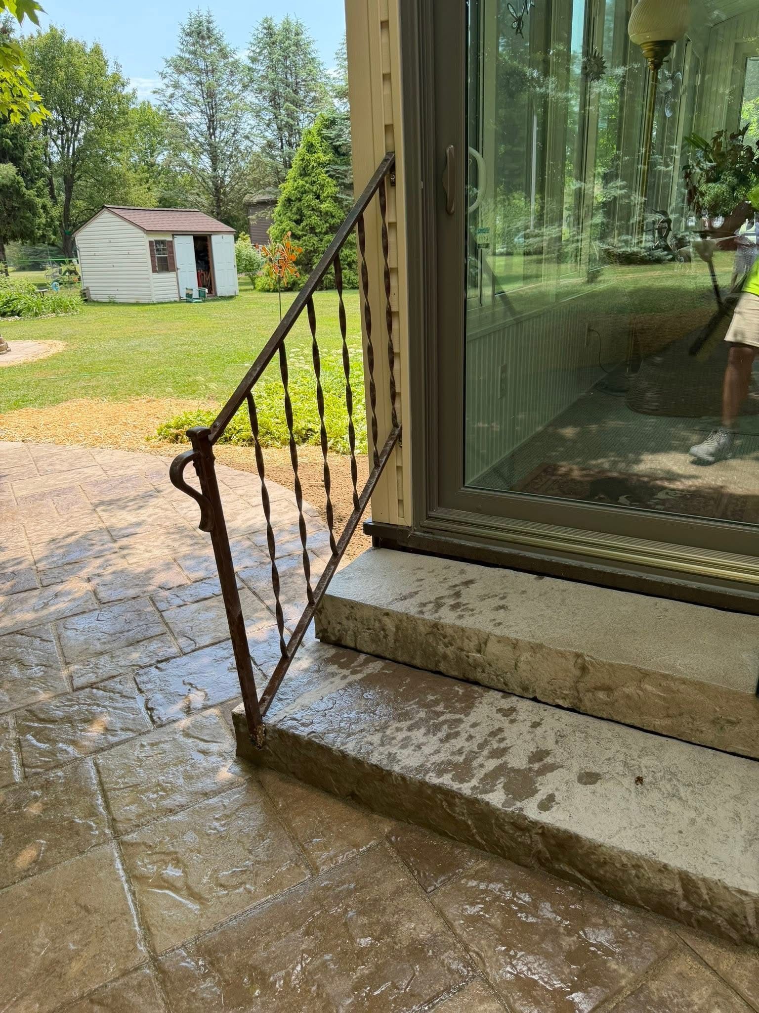 Stone patio beside sliding glass door and black railing, with a small shed and lawn in the background.
