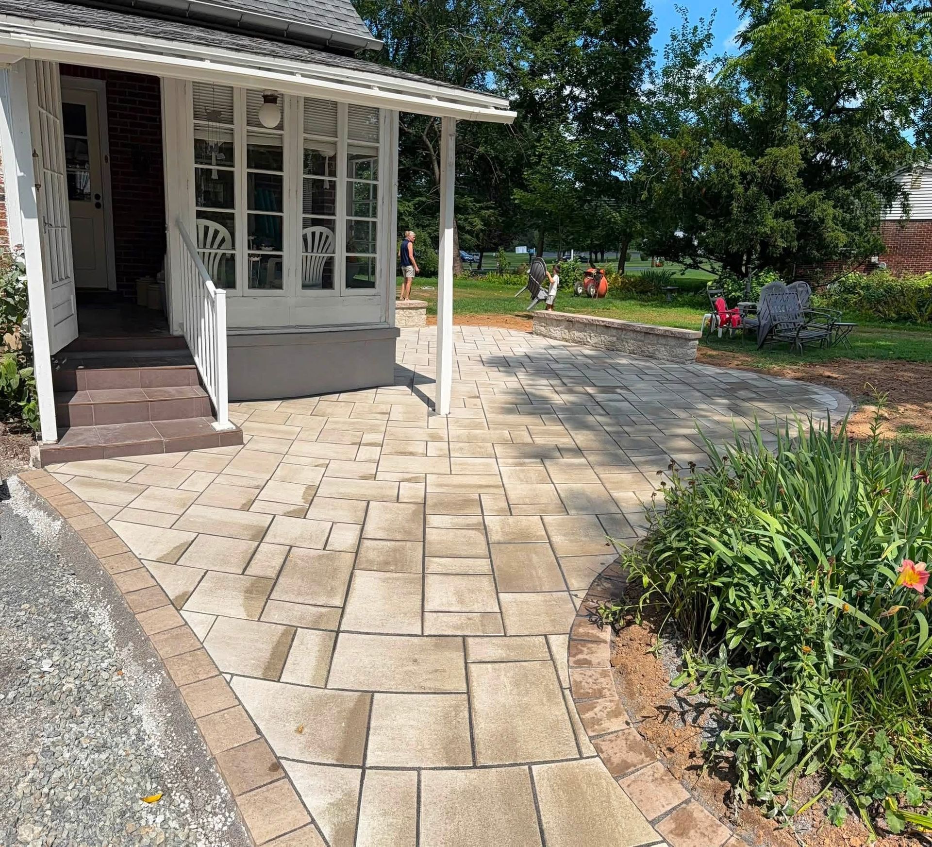 Brick patio beside a white house porch, with garden beds and trees in bright sunlight