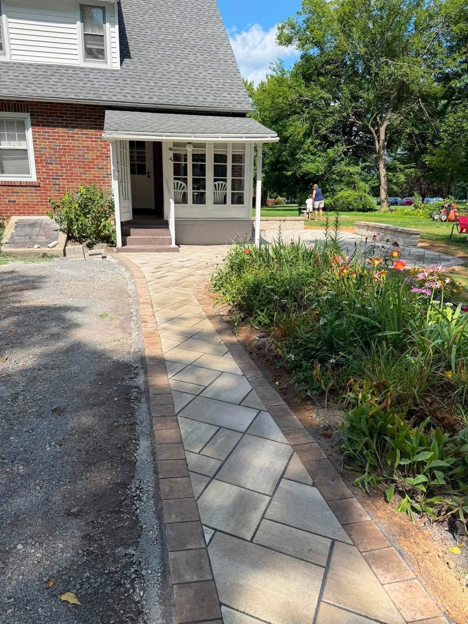 Brick house entrance with a curved stone walkway, gravel driveway, and flower bed in bright daylight