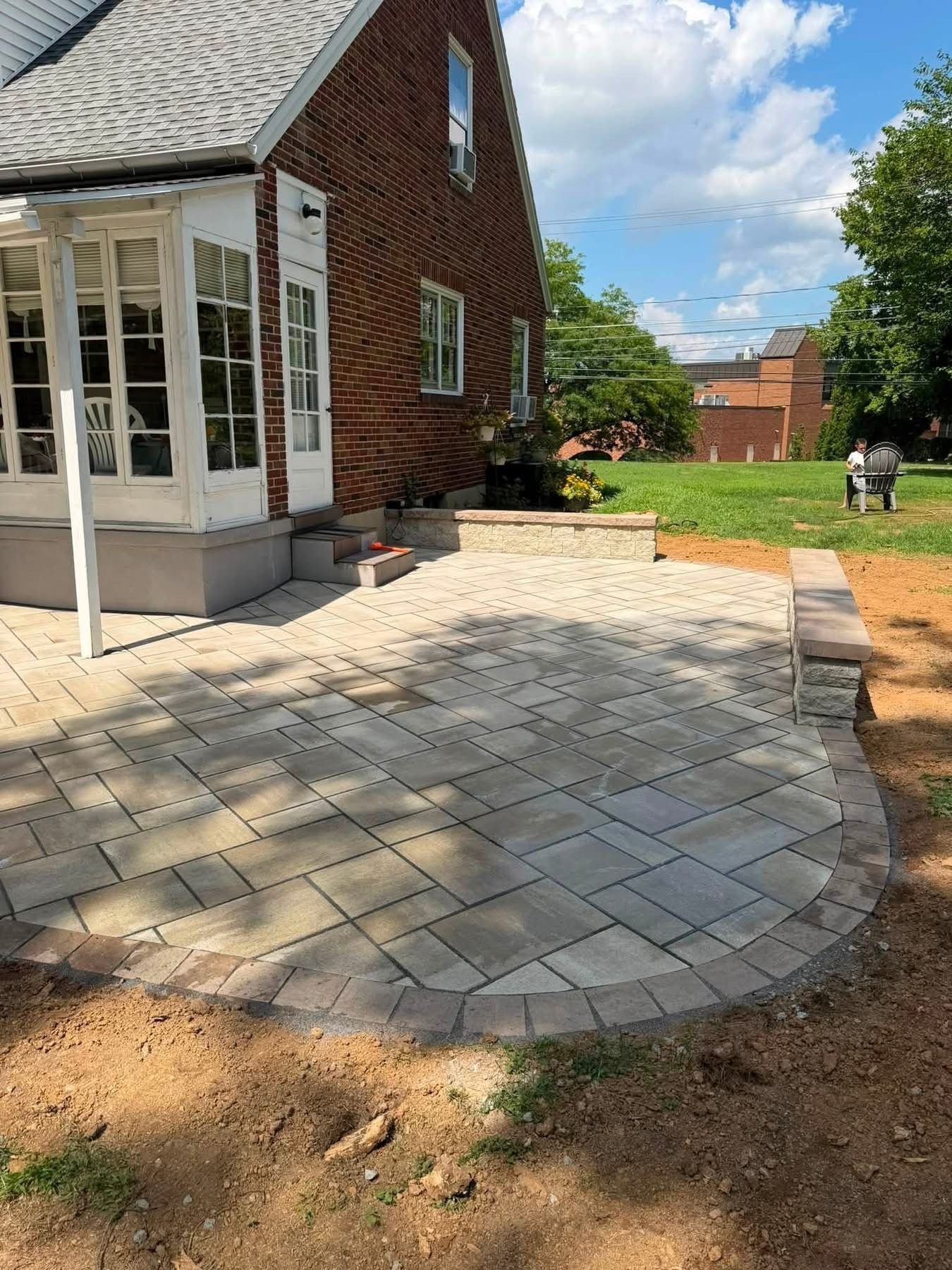 Brick house patio with newly laid pavers, a curved edge, and a grassy backyard under a blue sky.