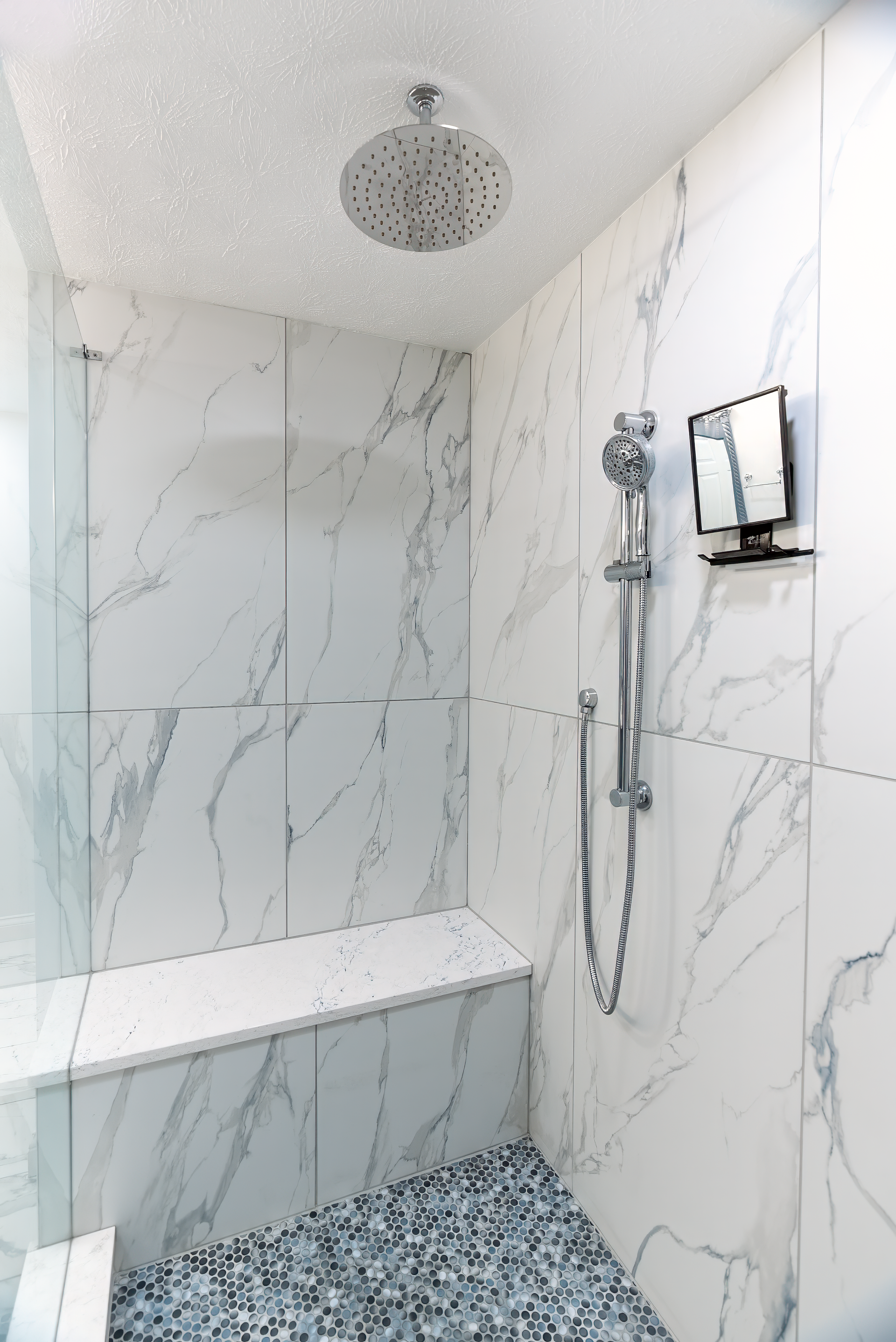 Shower with marble-look tile, pebble floor, built-in bench, and rain shower head. Handheld shower and TV are mounted.