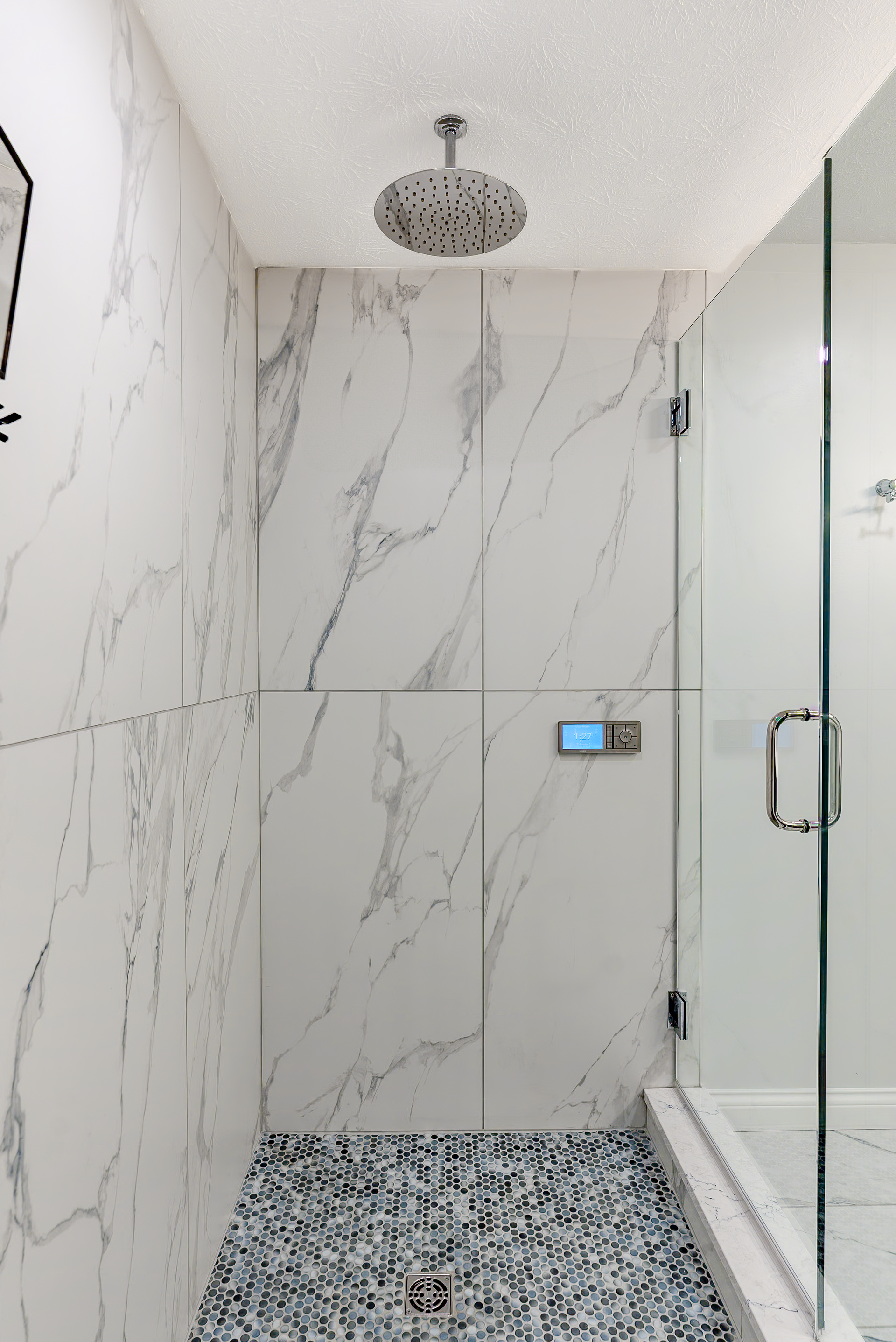 White marble-tiled shower with pebble floor, glass door, and overhead rain showerhead.