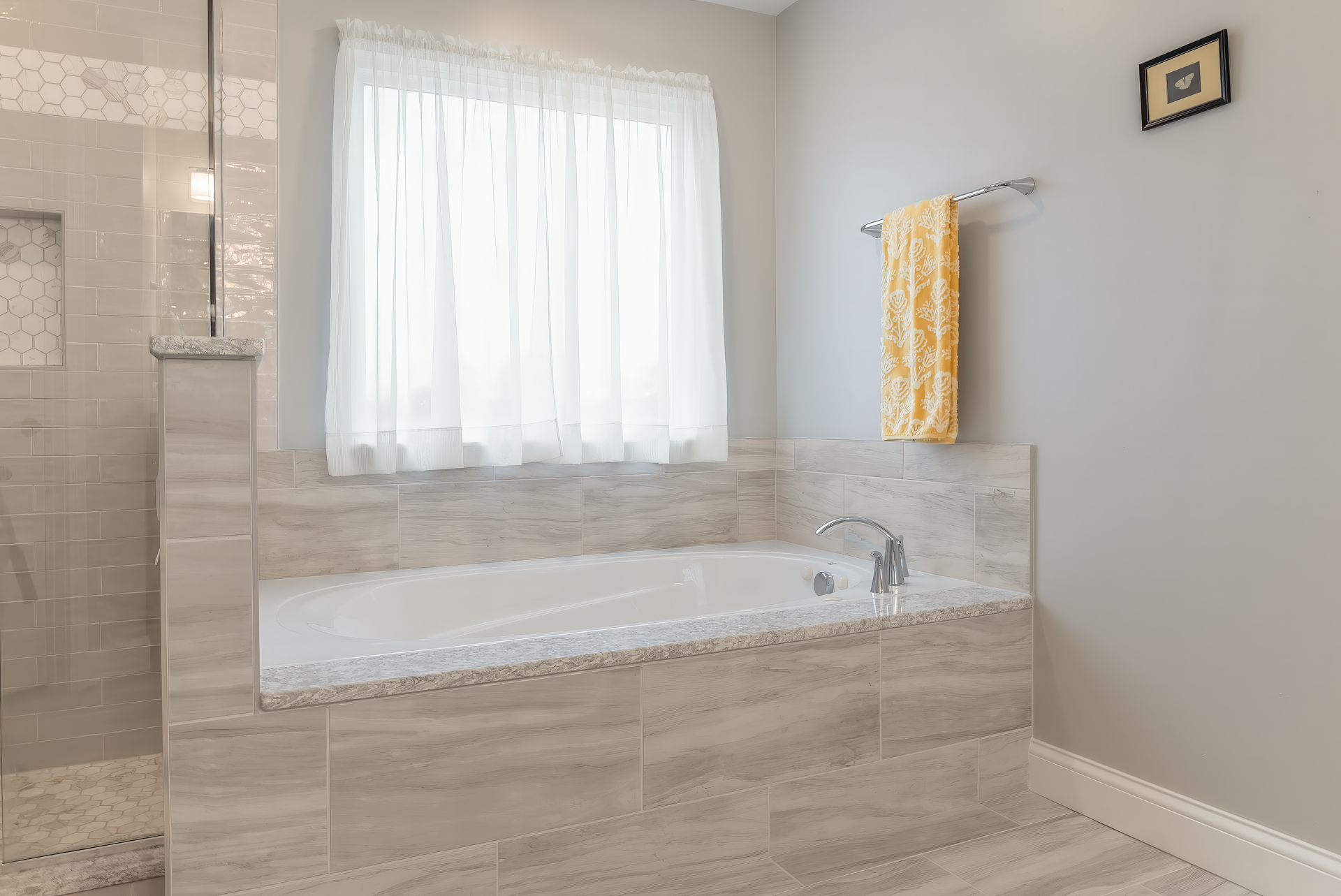 Bathroom with built-in tub, tiled surround, shower, sheer window curtain, and a yellow towel on a towel rack.