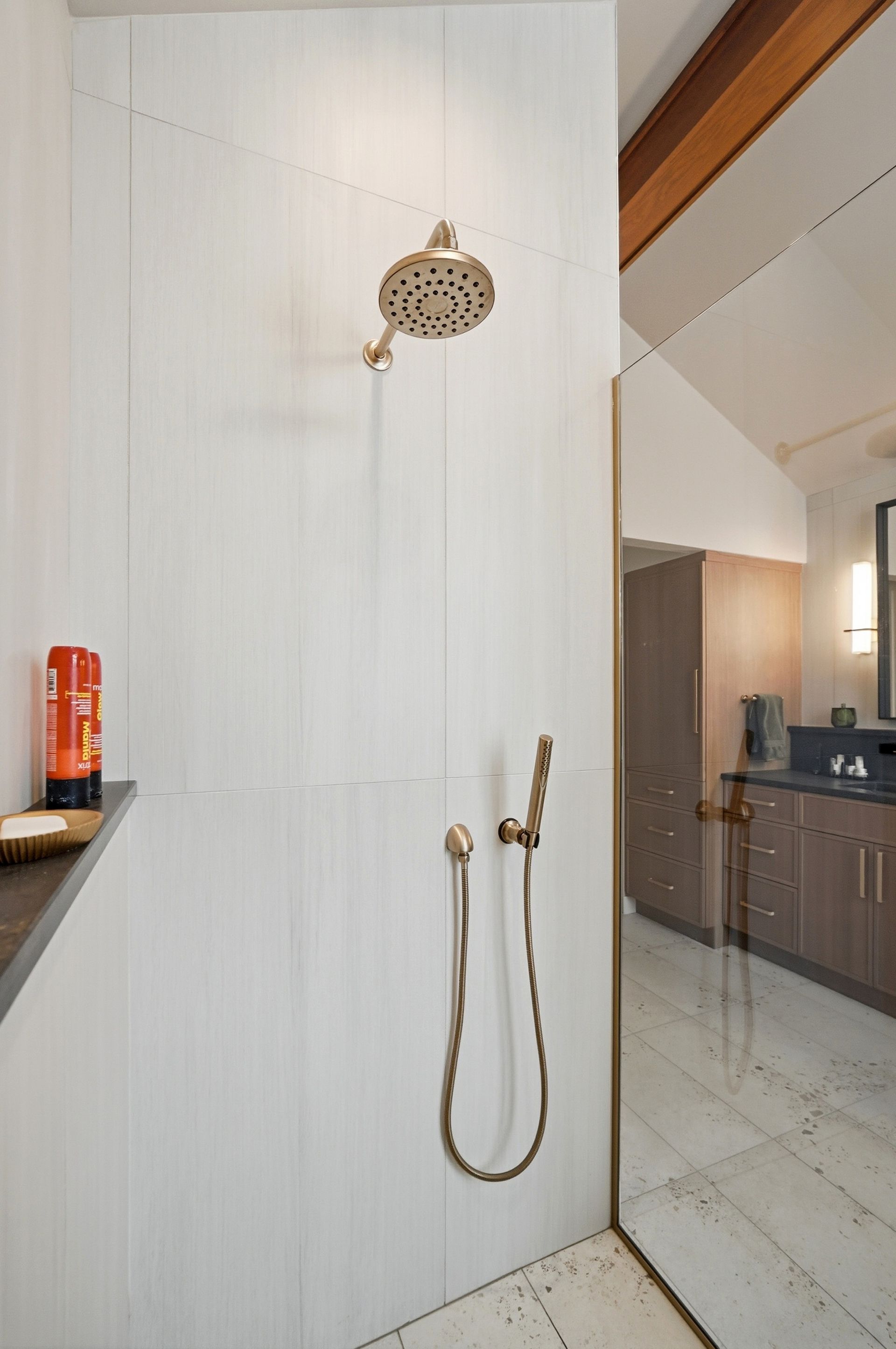Shower with bronze fixtures against white vertical paneling; mirror reflects bathroom with cabinetry.