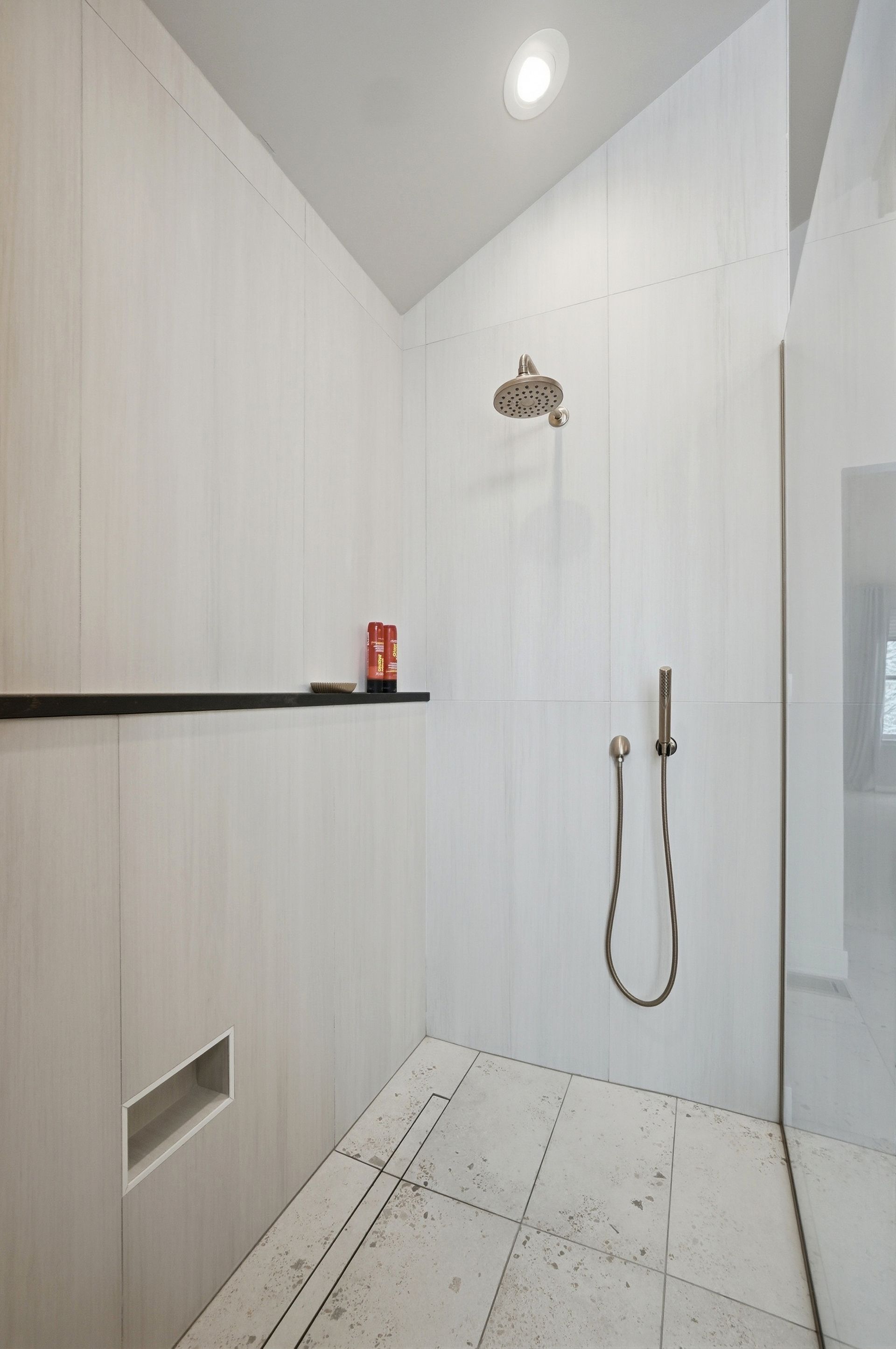 Modern shower with white tiled walls and floor, showerhead, handheld shower, and black shelf.