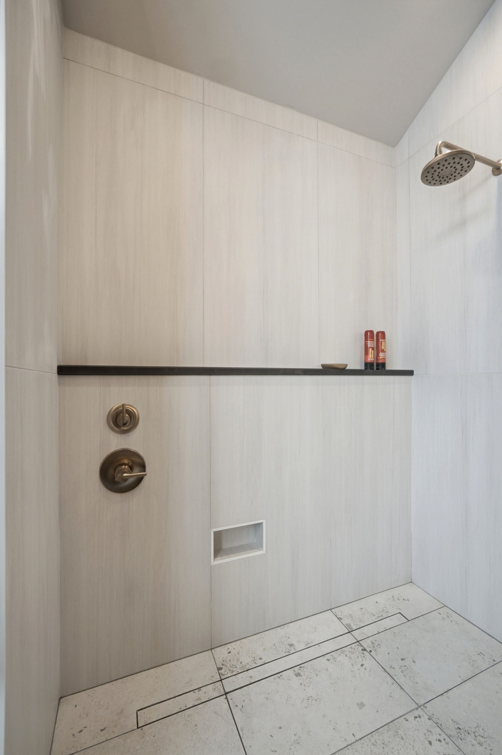 Shower stall with light wood-look tile, dark shelf, bronze fixtures, and rectangular drain in the floor.