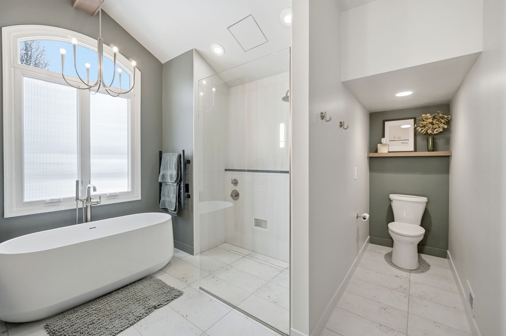 Modern, bright bathroom with white soaking tub, walk-in shower, and toilet area. Gray walls, light tile, and window.