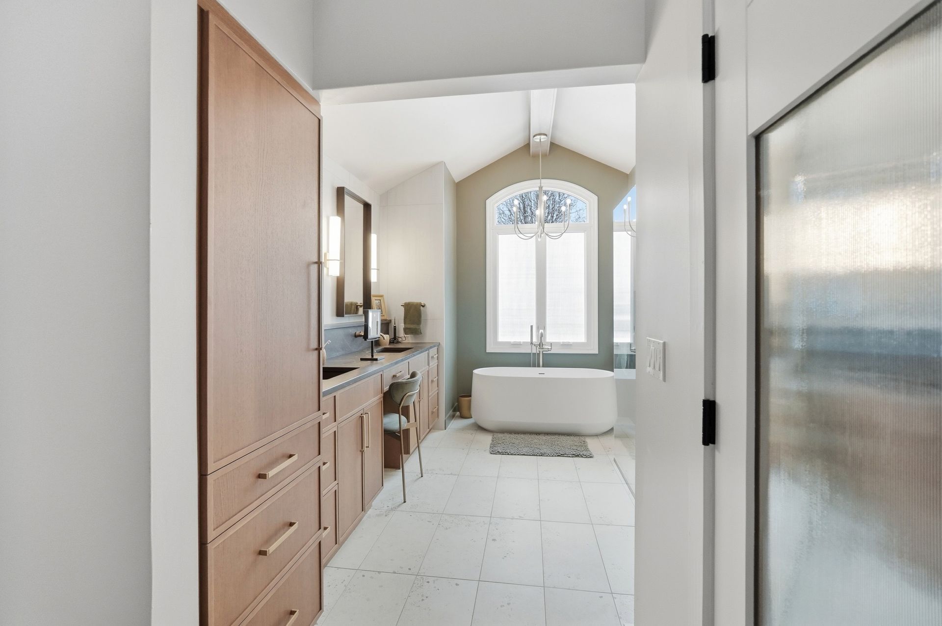 Bathroom interior: white walls and floor, wooden cabinet, bathtub near a window, open doorway.