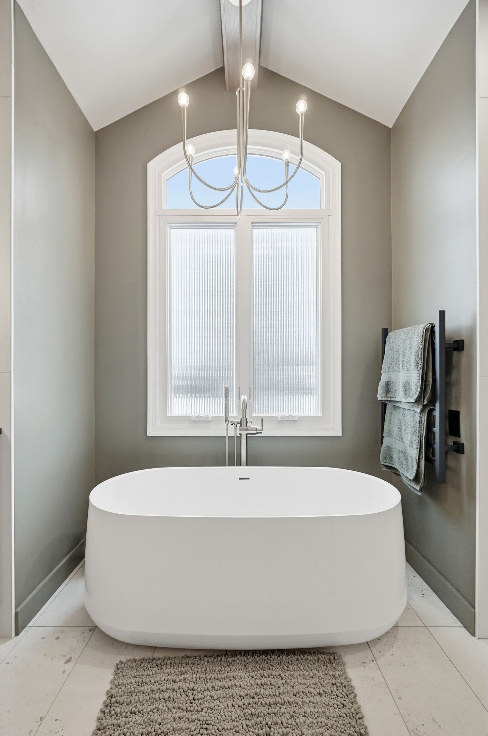 Modern bathroom with white tub, large window, and gray walls; a chandelier hangs overhead.