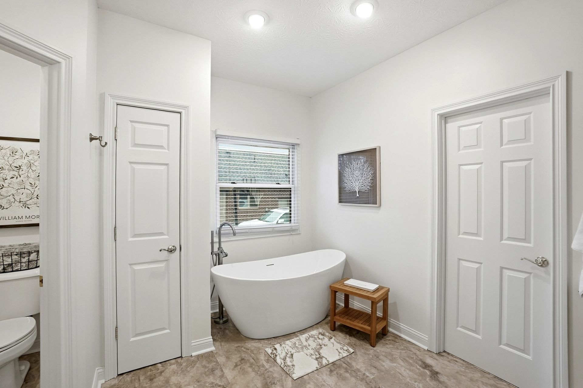 White bathroom with a freestanding tub, wood stool, and two closed doors.