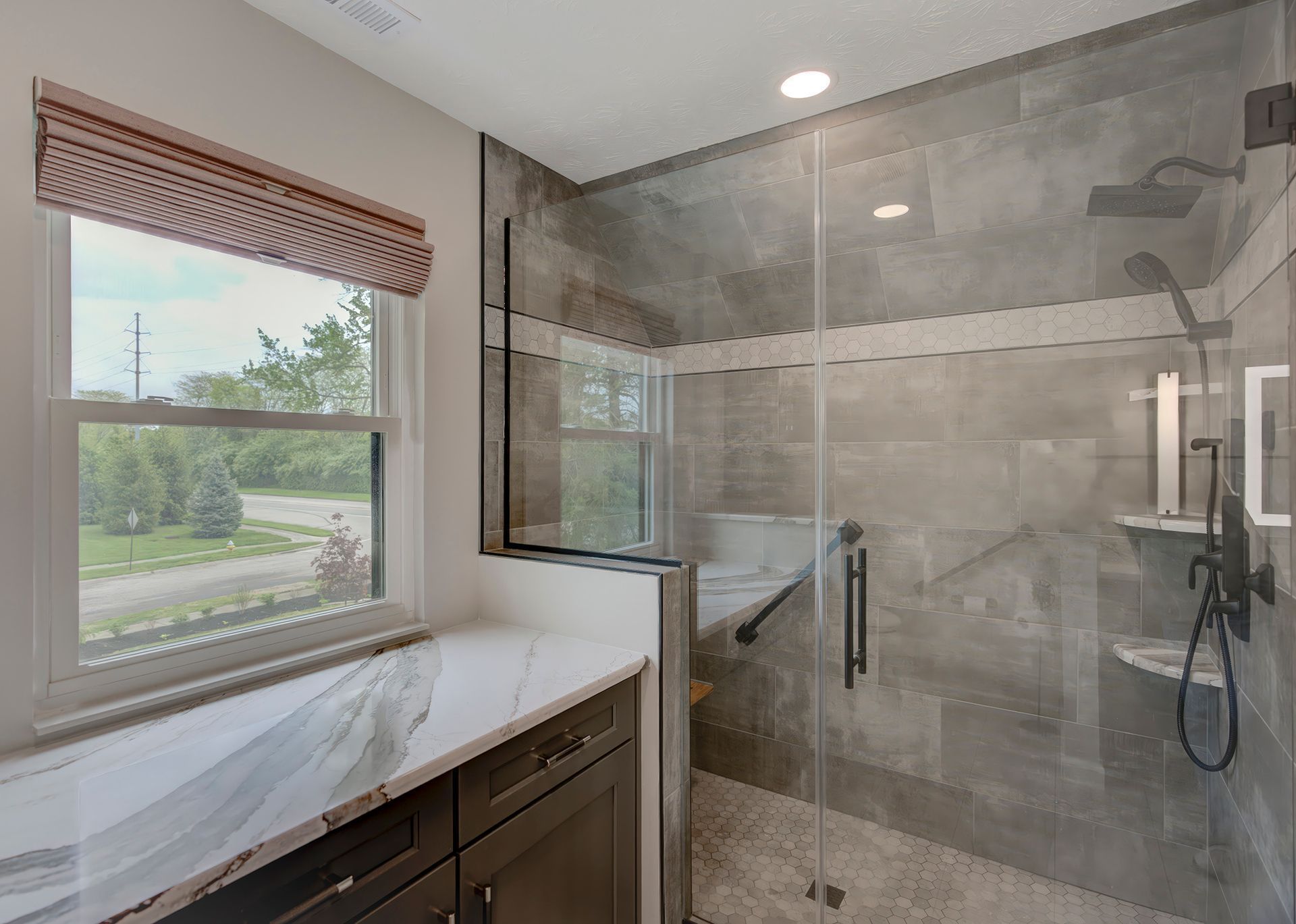 Modern bathroom with glass shower, gray tiles, and a window with a Roman shade.