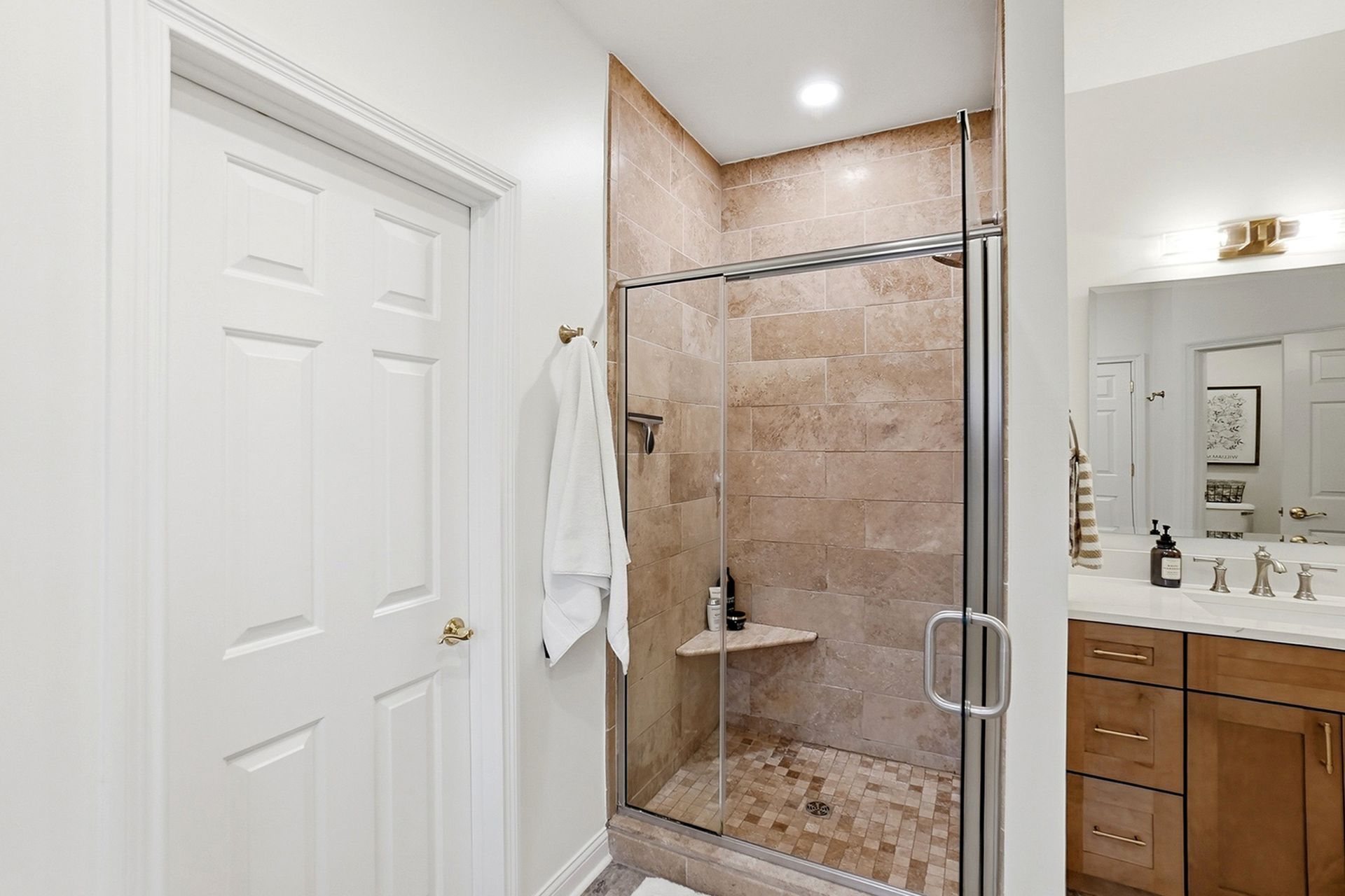 Bathroom with shower, tan tile, glass door, white robe, vanity, and closed door.