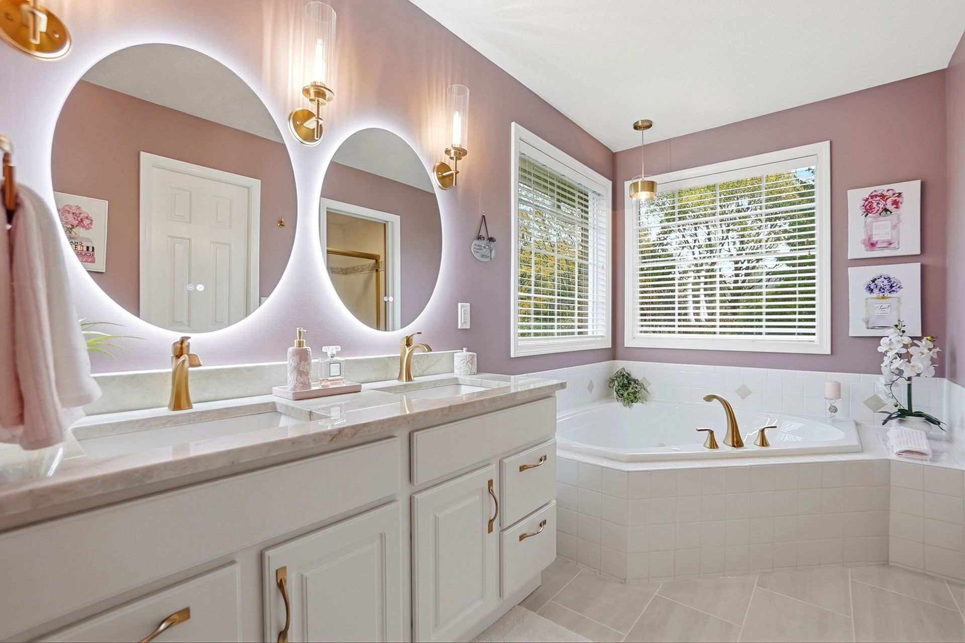 Bathroom with lilac walls, white cabinets, gold fixtures, round mirrors with lights, and a white bathtub.