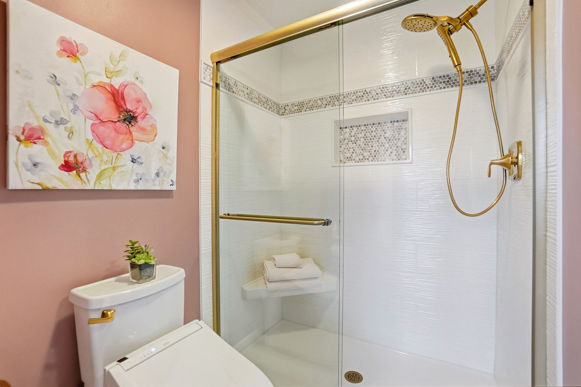 Bathroom with pink walls, gold shower fixtures, and floral artwork. Toilet in the foreground.