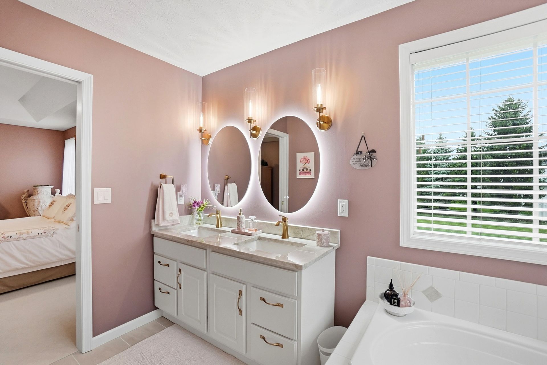 Bathroom with pink walls, white vanity, round mirrors with lights, and window.