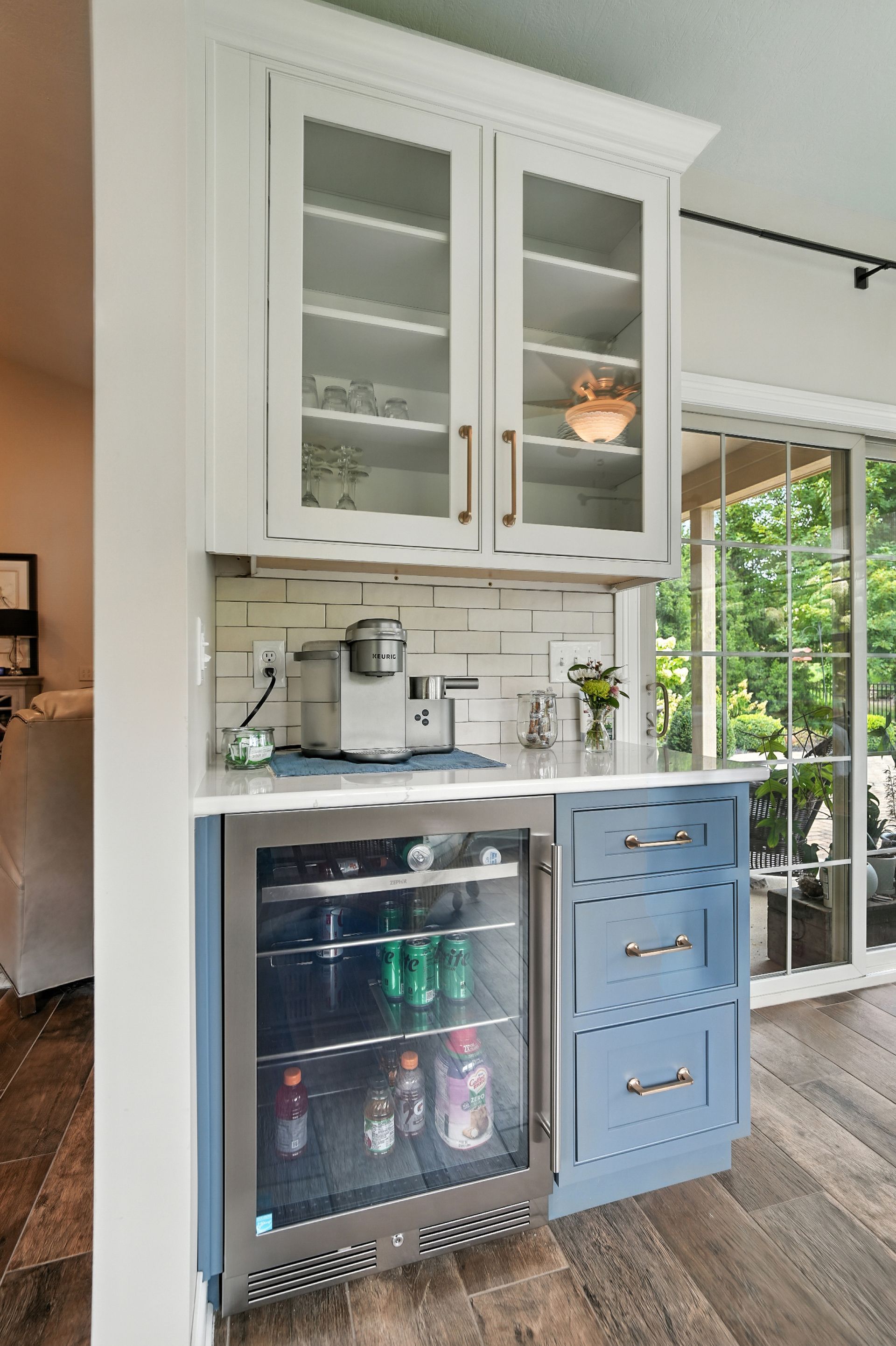 Built-in coffee station: white cabinets, glass doors, blue base with fridge and drawers, overlooking a patio.