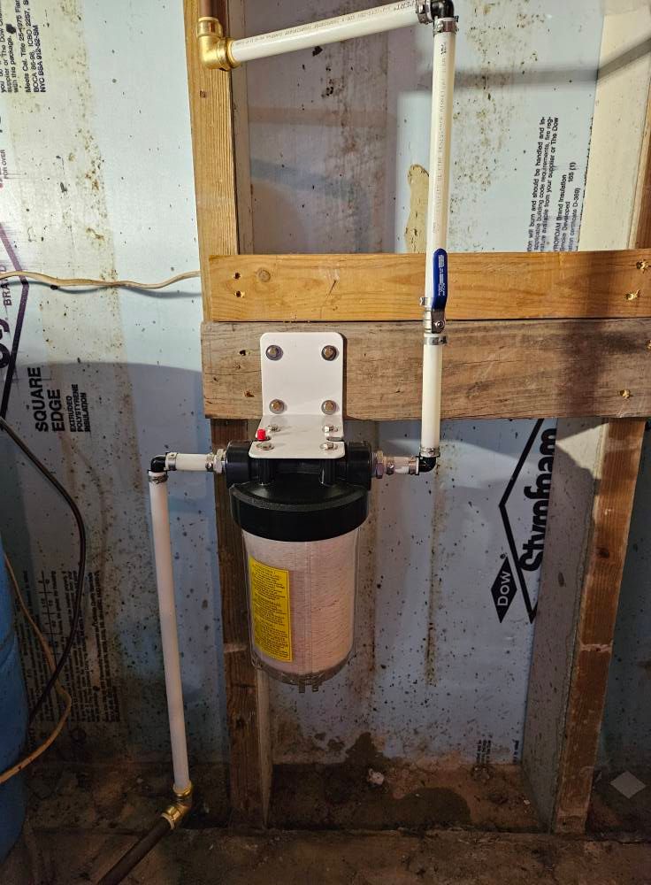 Water filter system with white pipes and black canister, mounted on wooden frame against wall.