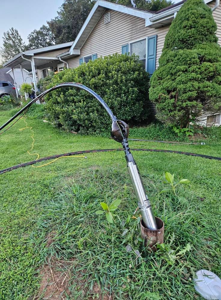 Well pump with black hoses in a grassy yard, near a house with bushes and a tree.