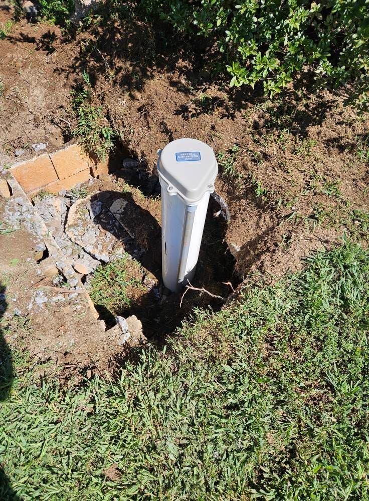 White well casing with a gray lid in a hole dug in the ground, near a brick wall and grass.