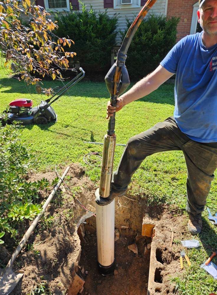 Man installing a well pump in a yard. He's holding the pump, which extends into a white pipe in the hole.