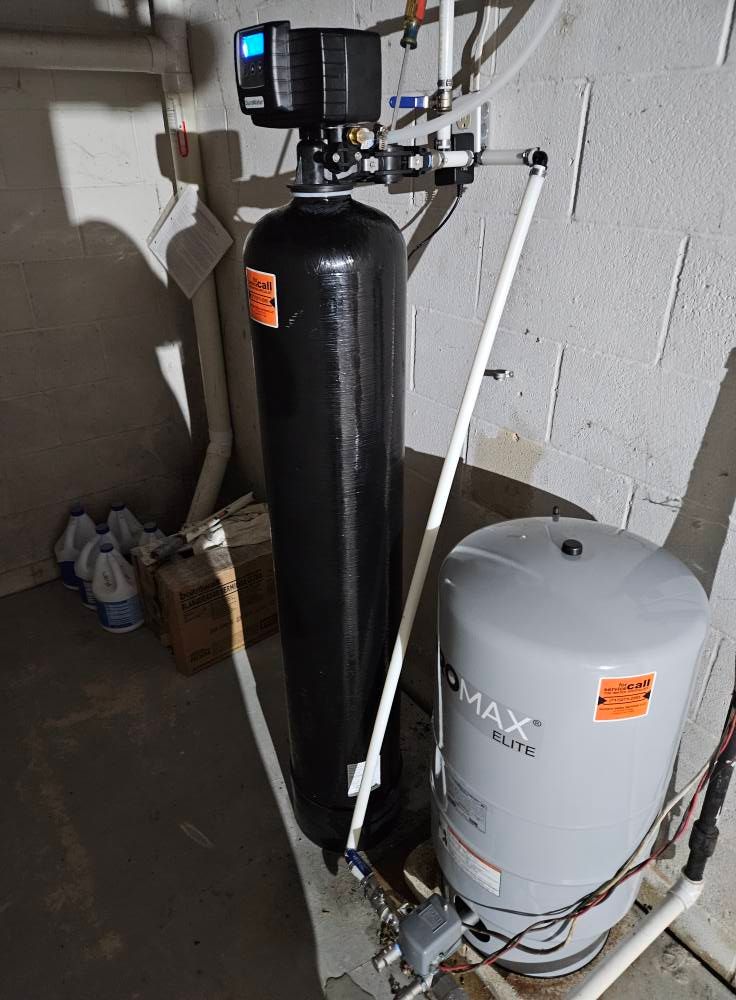 A water filtration system in a basement. It has a tall black tank, digital control panel, and a smaller gray tank.