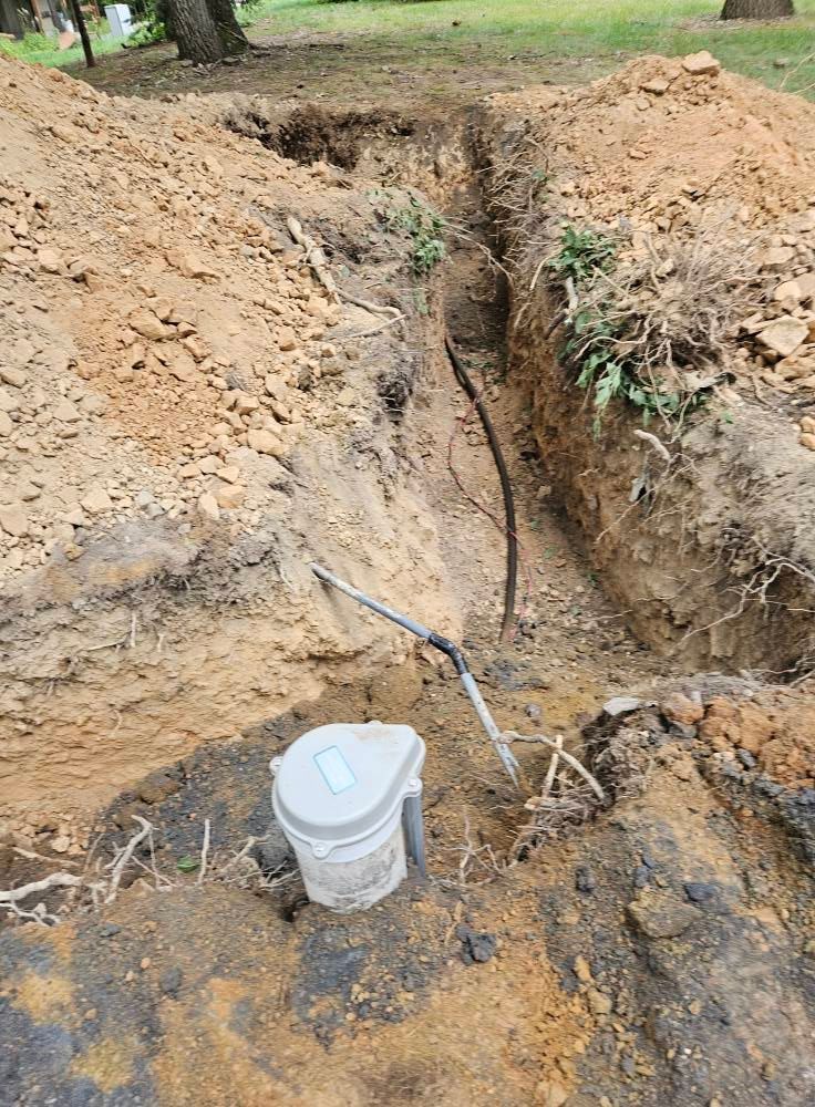 Trench dug in dirt with electrical wiring, and a gray device at the edge.