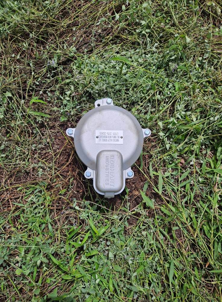 Silver utility box on the ground, surrounded by green grass and weeds.
