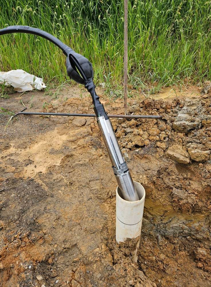 A submersible pump being lowered into a white PVC well casing in a dirt ground, with a black hose attached.