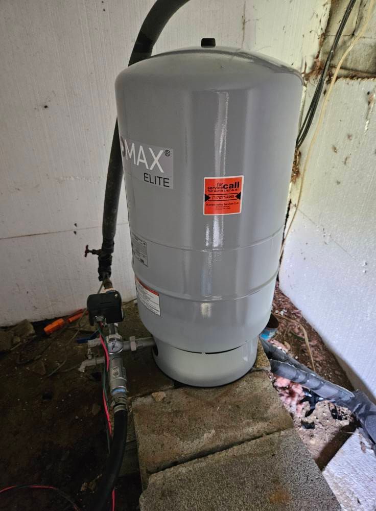 A gray water pressure tank sits on concrete blocks indoors near pipes and wiring.