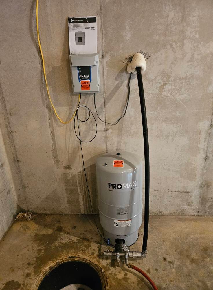 Sump pump system in a concrete-walled basement. Gray tank, electrical components, and piping are visible.
