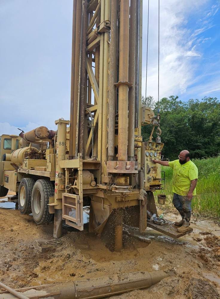 A bearded person in a green shirt stands beside a drilling rig, muddy ground, overcast sky.