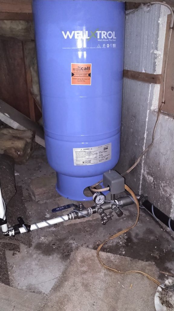 Blue well water tank in a basement, with associated pipes and pressure control devices.