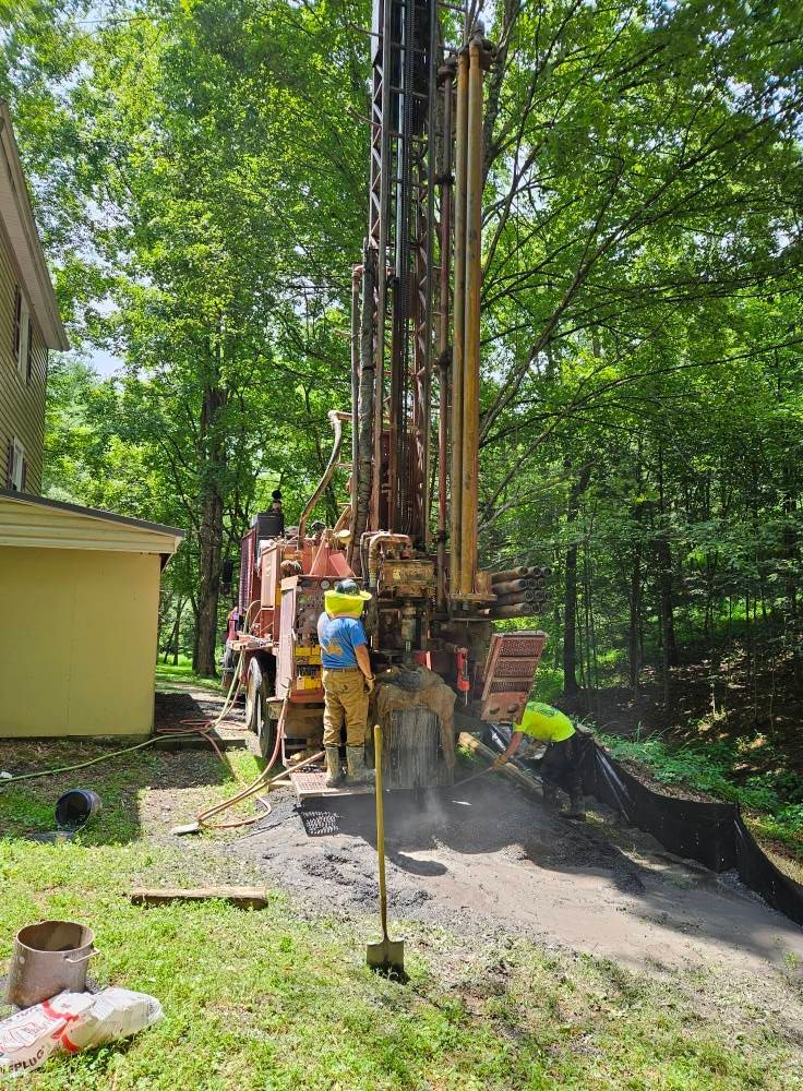 Drilling rig with workers near a house in a wooded area.  One worker in yellow hard hat.