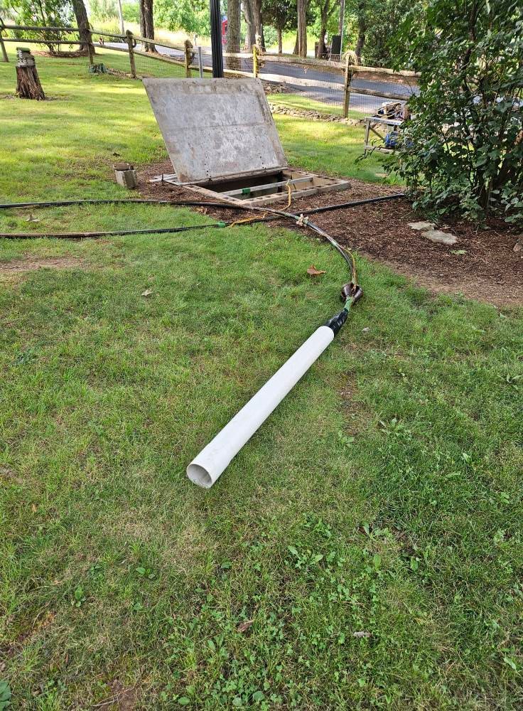 A white pipe on grass leads to an open underground utility access point. Cables and trees are in the background.