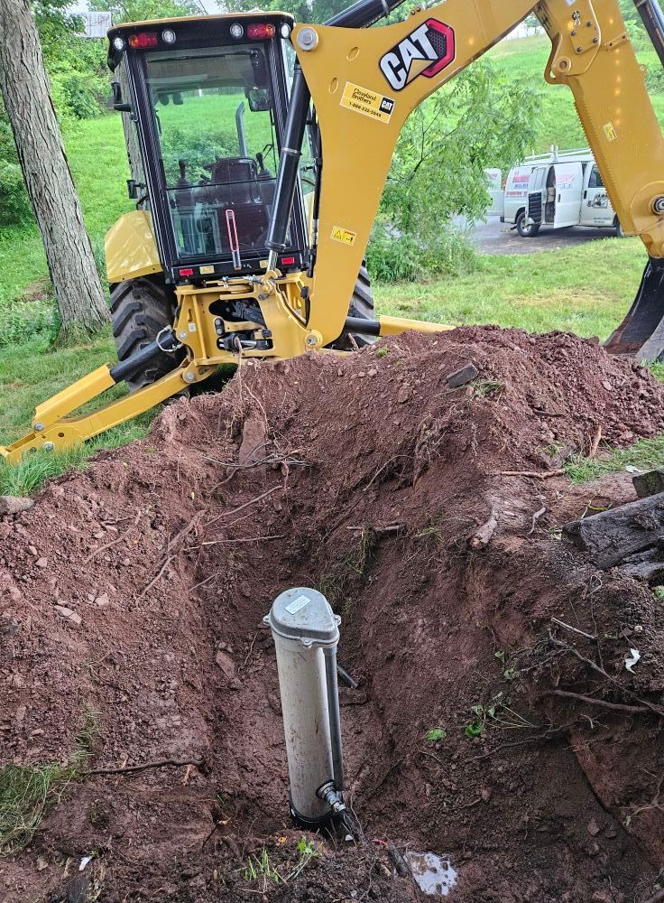 Yellow CAT excavator working on a dirt trench around a cylindrical object in an outdoor setting.
