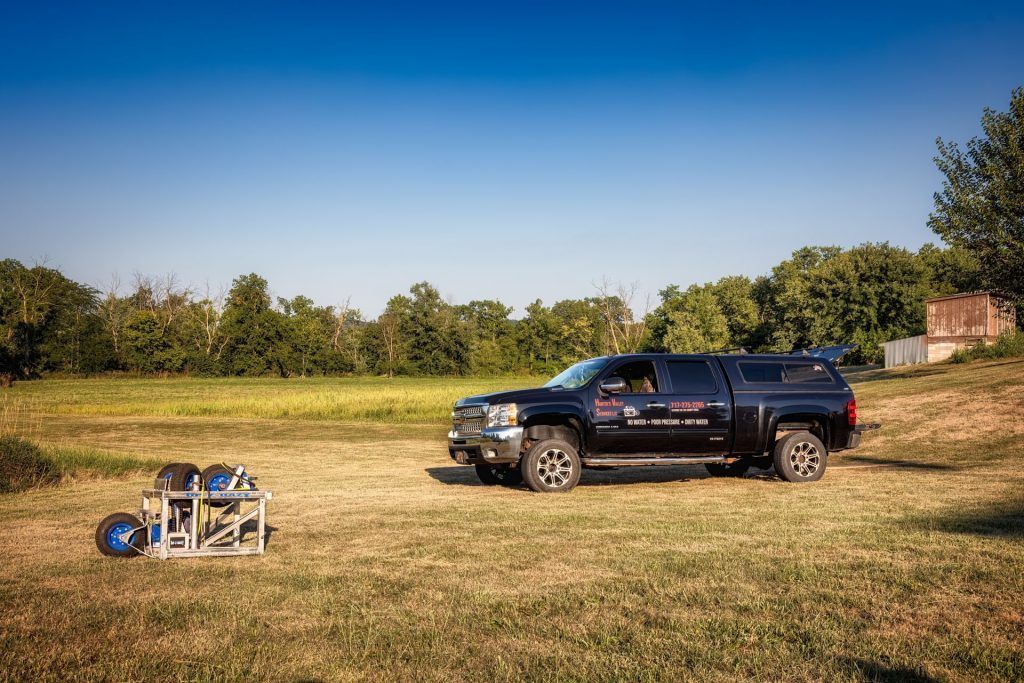 Black truck parked in a grassy field next to a small, silver mechanical device; trees and blue sky in background.