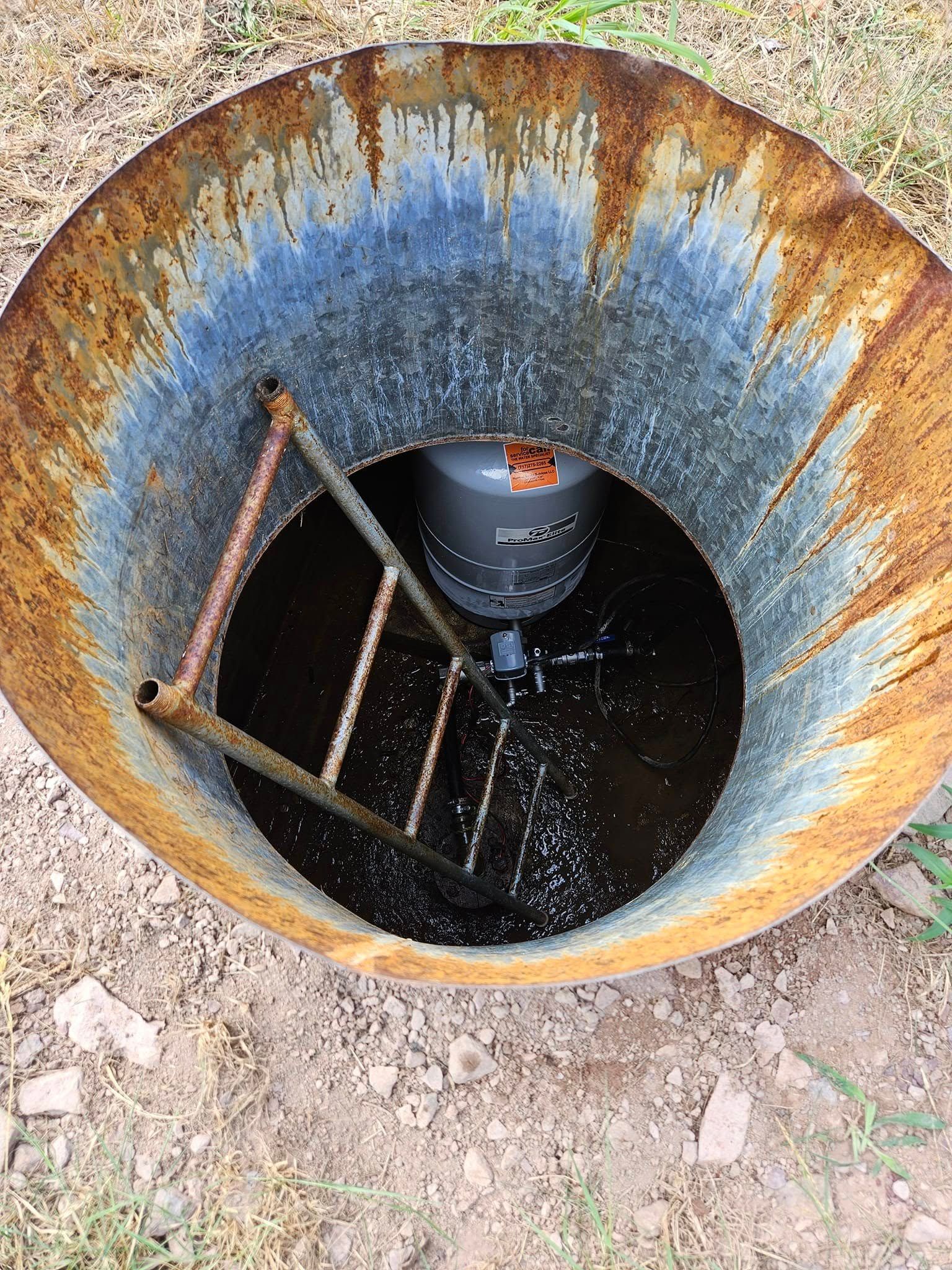 Rusty metal drum with dark liquid, a cylindrical object, and a metal ladder-like structure inside.